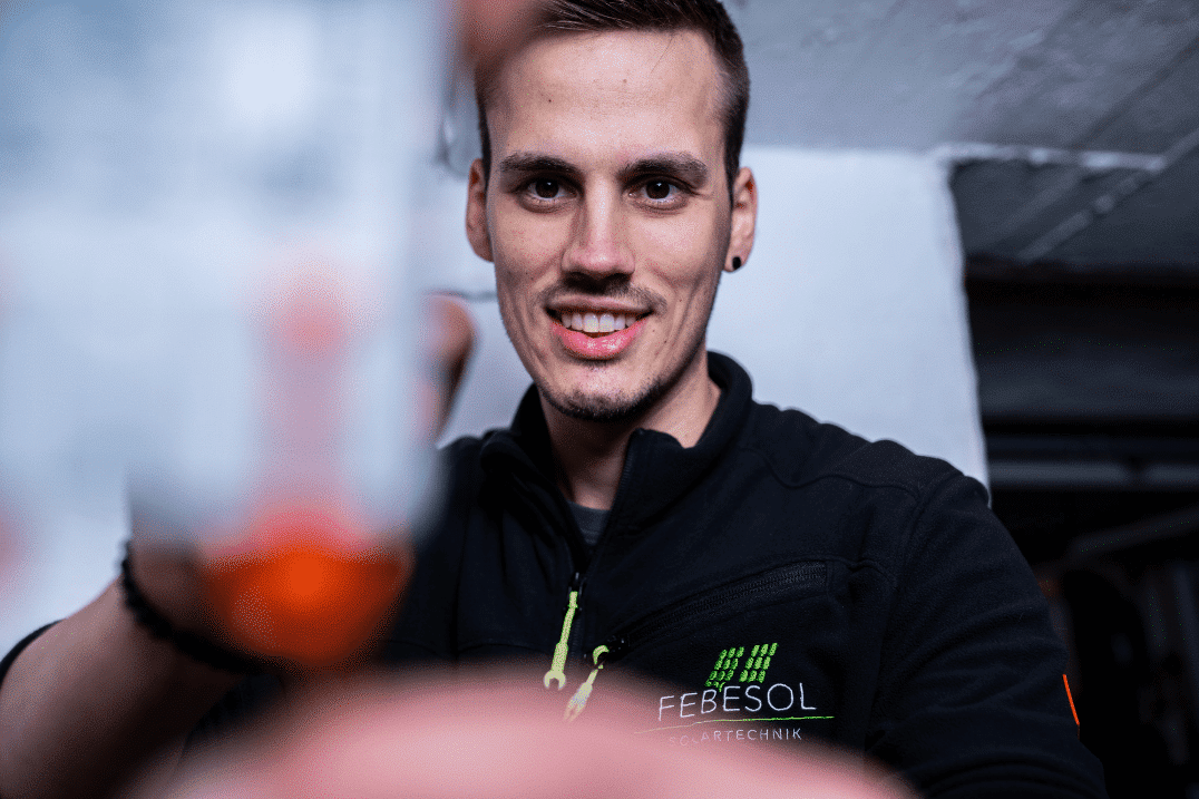 Ein Mann lächelt, während er eine Flasche Flüssigkeit vor einer Solaranlage hält.