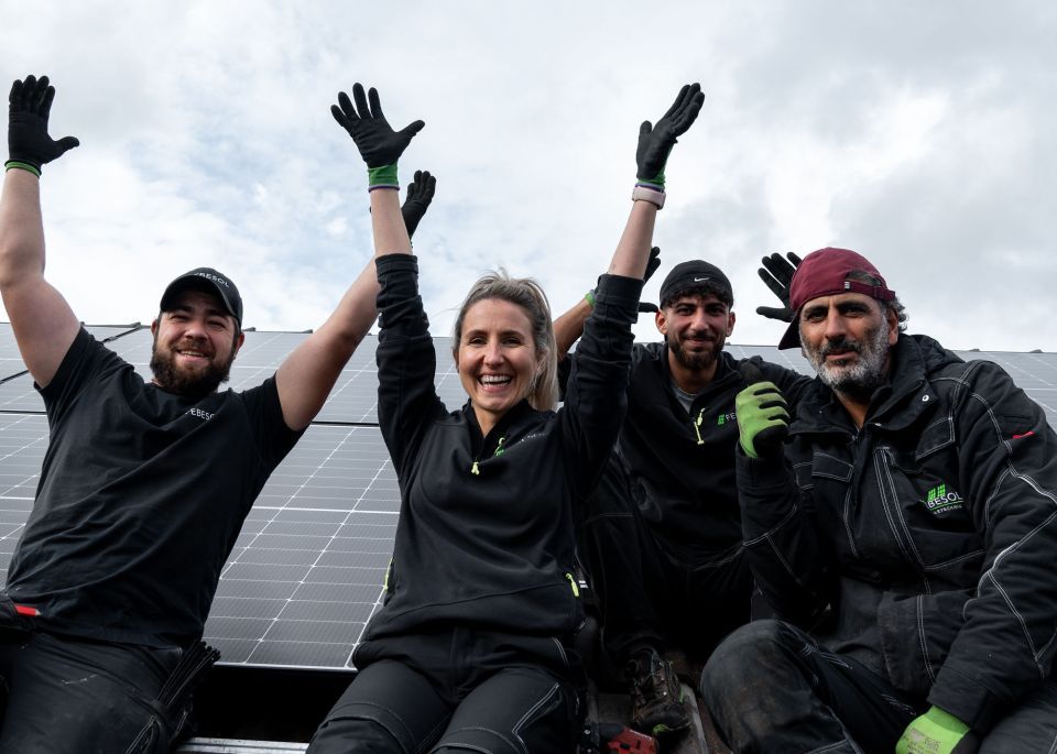 Ein Team von vier Arbeitern in schwarzen Uniformen mit grünen Handschuhen hebt fröhlich die Hände auf einem Dach der Solaranlage Ludwigshafen.