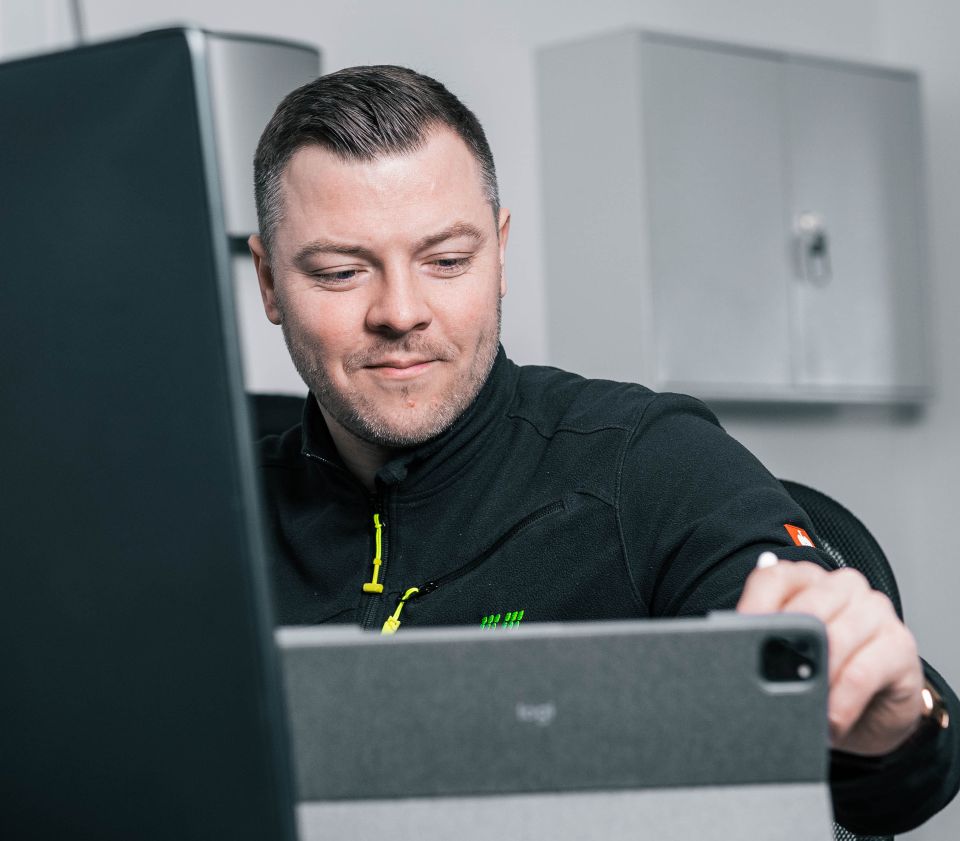 Ein Mann in einem schwarzen Hemd mit einem grünen Logo passt in einer modernen Büroumgebung die Einstellungen eines Laptops an.
