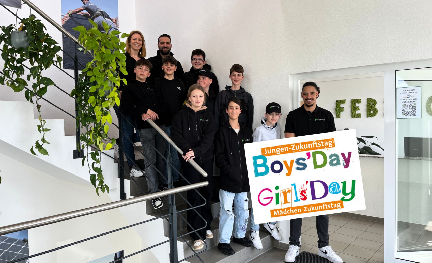 Eine Gruppe von Jungen und Mädchen sowie zwei Erwachsene stehen auf einer Treppe und halten ein buntes Schild mit der Aufschrift „Girls‘ & Boys‘ Day bei FEBESOL“. Die Szene scheint sich in einem Innenbereich in der Nähe einer Pflanze zu befinden.
