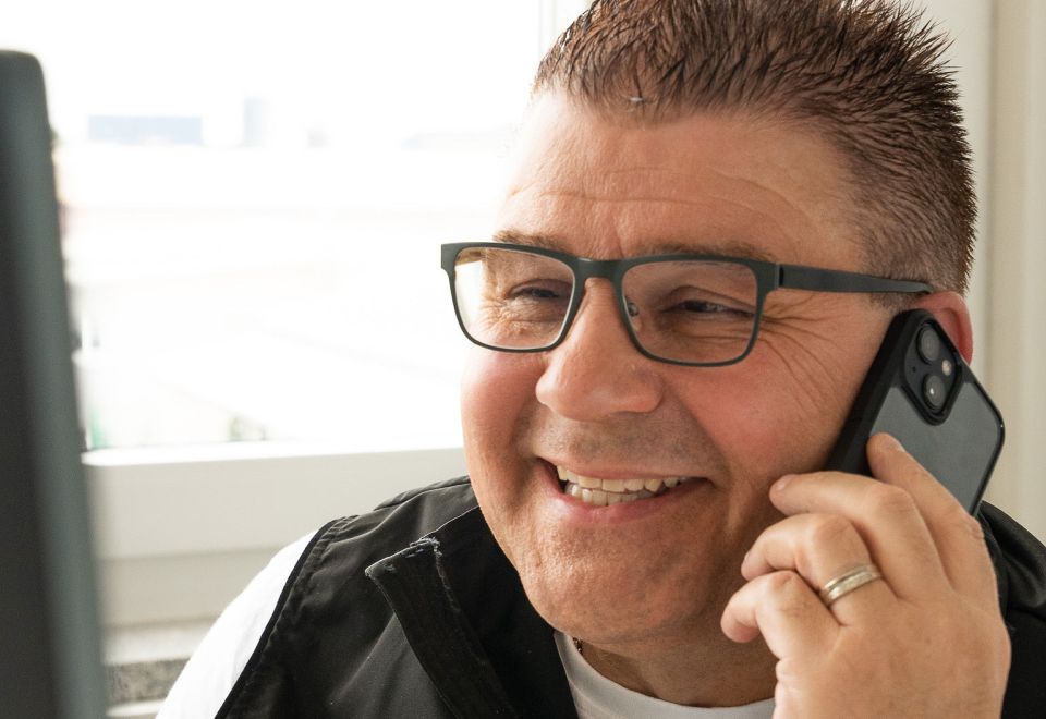 Ein lächelnder Mann mit Brille telefoniert in einem Innenbereich mit einem Smartphone.
