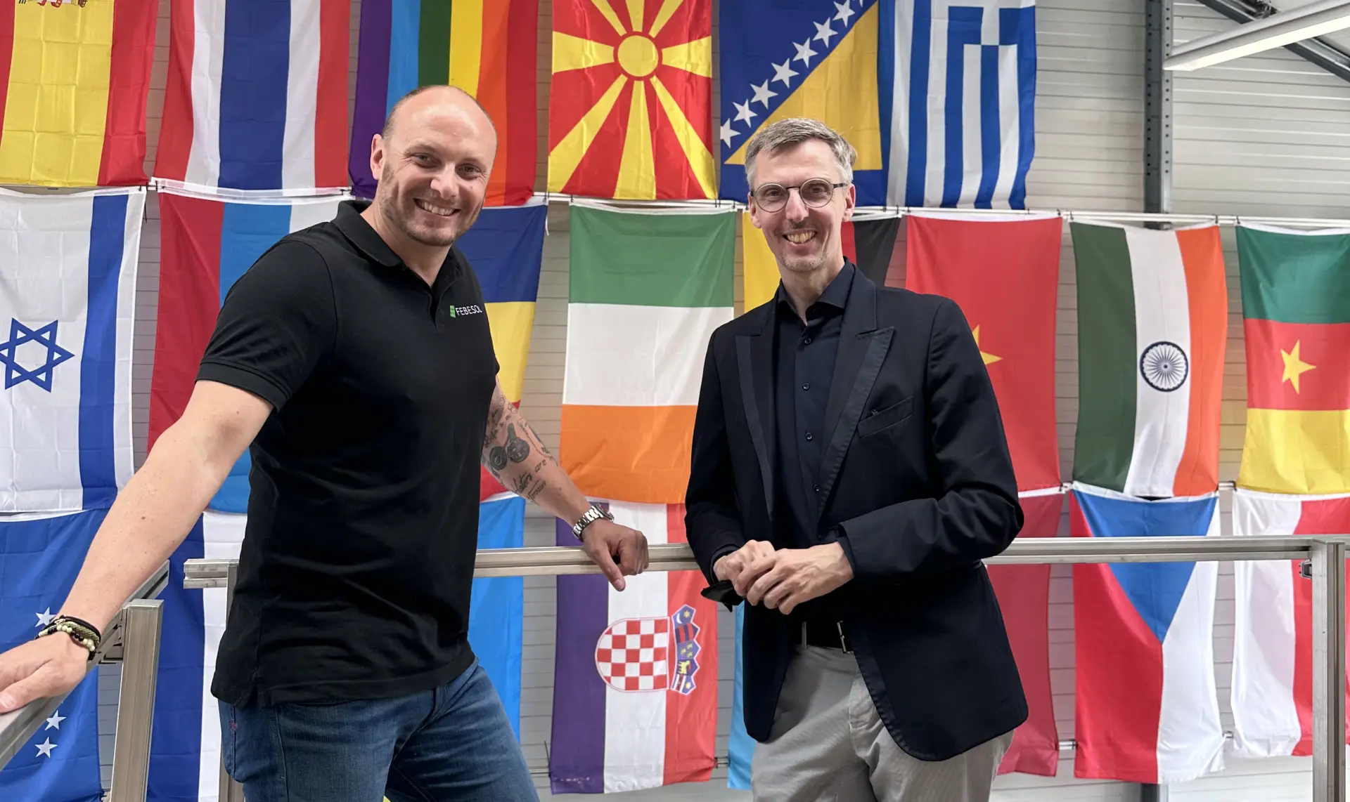 Zwei Männer schütteln sich vor einer Ausstellung verschiedener internationaler Flaggen die Hände bei der Girls‘ & Boys‘ Day-Veranstaltung bei FEBESOL in einem Innenbereich.