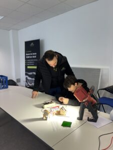 Erwachsener hilft einem Kind an einem Tisch bei einem Elektronikprojekt, im Hintergrund sind pädagogische Febesol-Banner zu sehen.
