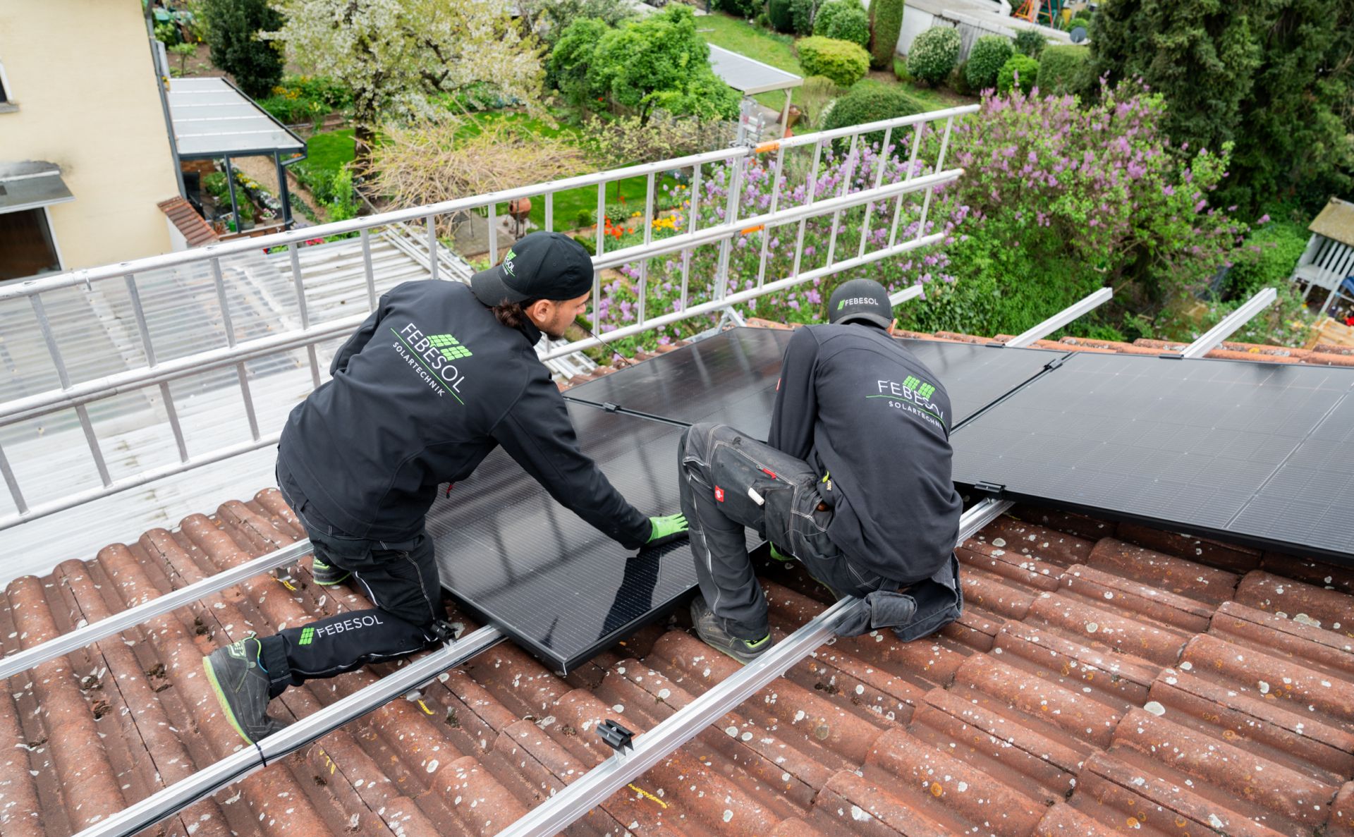 Zwei Techniker in schwarzen Jacken installieren mit Unterstützung des KFW-Förderprogramms 442 Solarmodule auf einem Ziegeldach vor einer grünen Wohnkulisse.