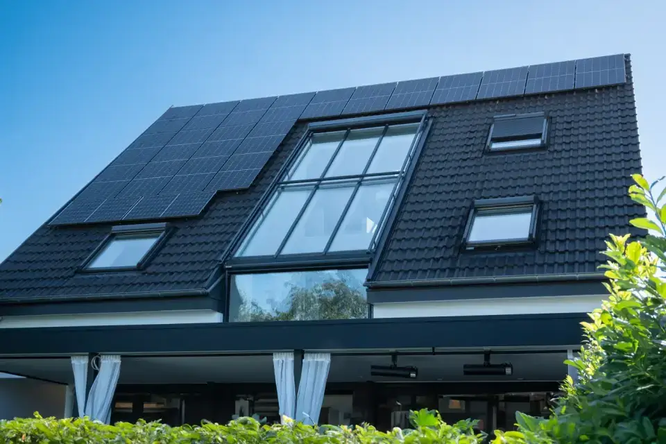 Modernes Haus mit Solarmodulen auf dunklem Schrägdach, großem Mittelfenster, umgeben von Grün.