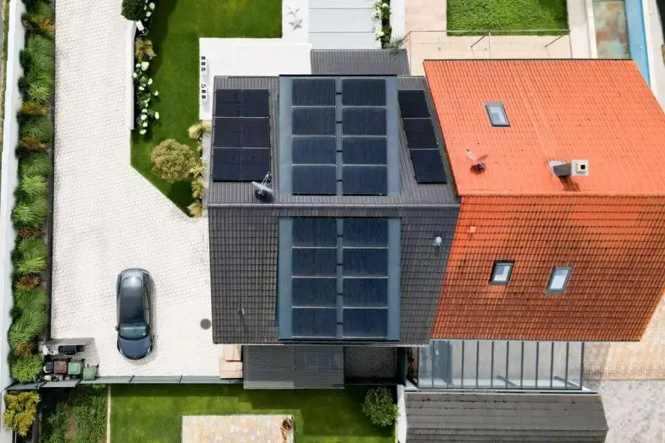 Luftaufnahme eines Hauses mit Solarmodulen auf dem Dach, Einfahrt und Garten mit Sträuchern und einem geparkten Auto.