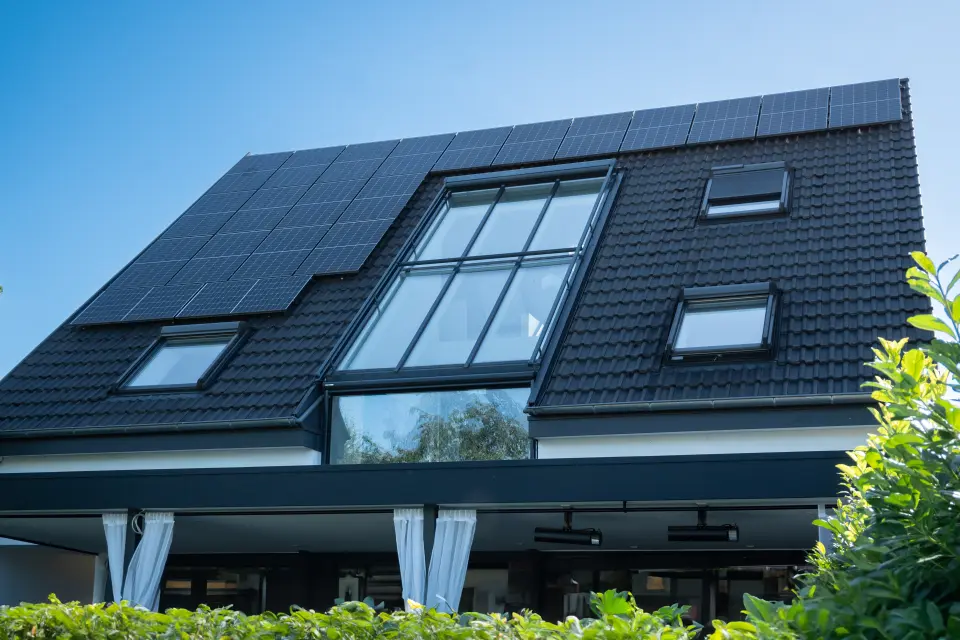 Ein modernes Haus mit schwarzen Dachziegeln und mehreren Solarmodulen, mit großen Fenstern, umgeben von Grün unter einem klaren blauen Himmel.