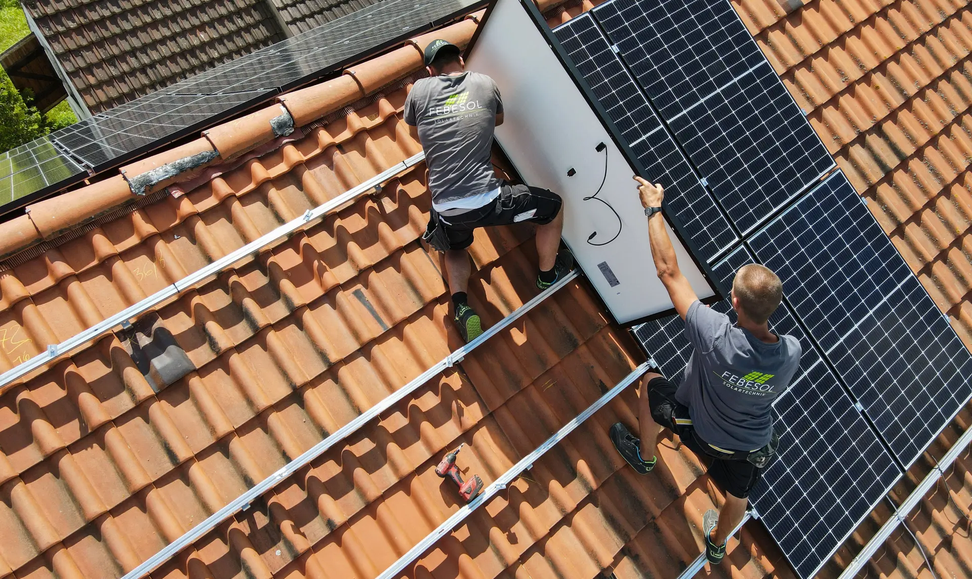 Zwei Arbeiter in grauen Hemden installieren systematisch eine 10 kWp PV-Anlage mit Speicher auf einem roten Ziegeldach. Einer hält ein Solarmodul, der andere befestigt es.