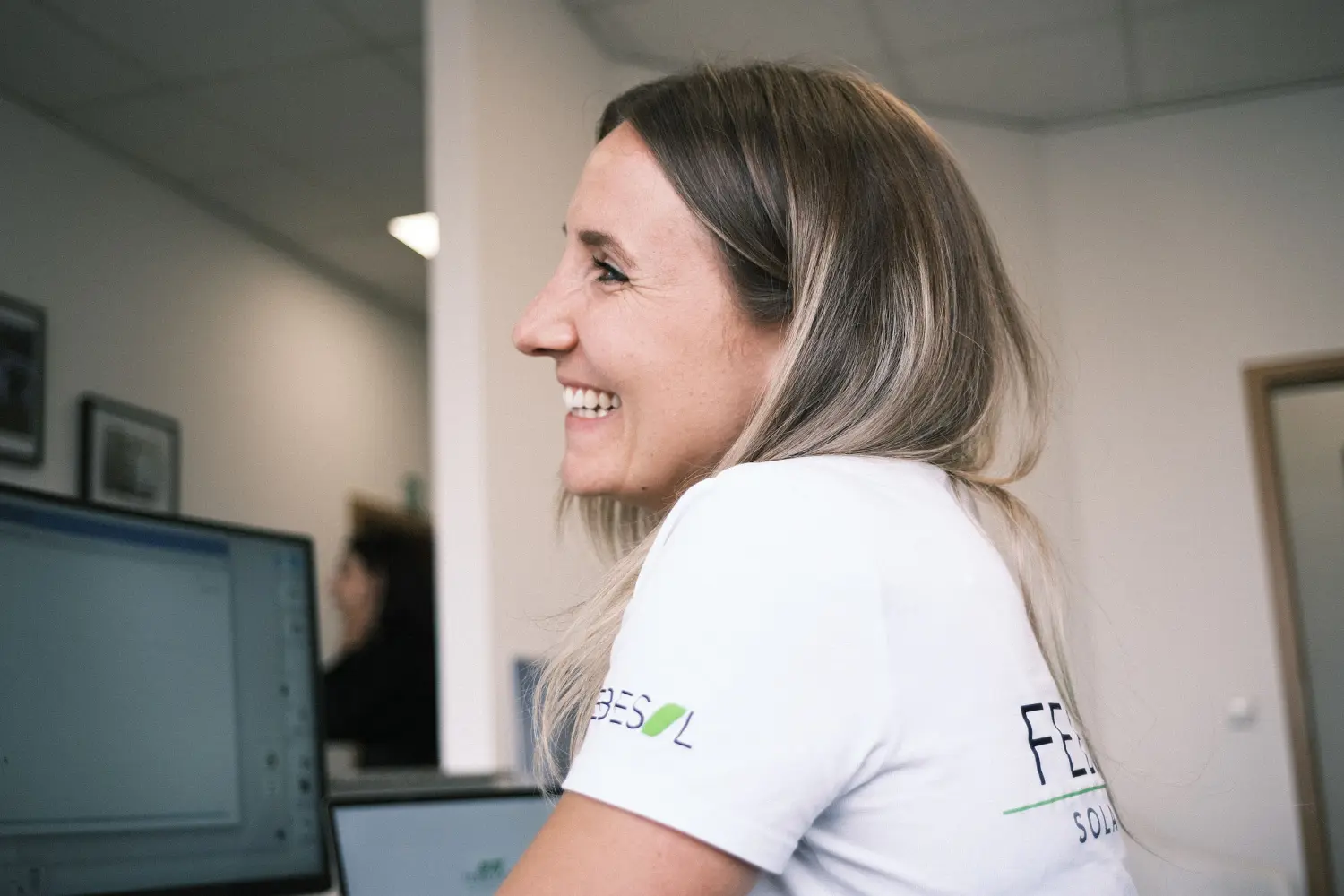 Eine Frau mit langen blonden Haaren lächelt, während sie in einem Büro auf einen Computerbildschirm schaut. Sie trägt ein weißes Hemd.