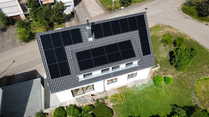 Luftaufnahme eines Hauses mit Sonnenkollektoren auf dem Dach, umgeben von Grünflächen und Straßen.