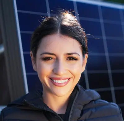 Eine lächelnde Person in einer schwarzen Jacke steht vor Solarmodulen.