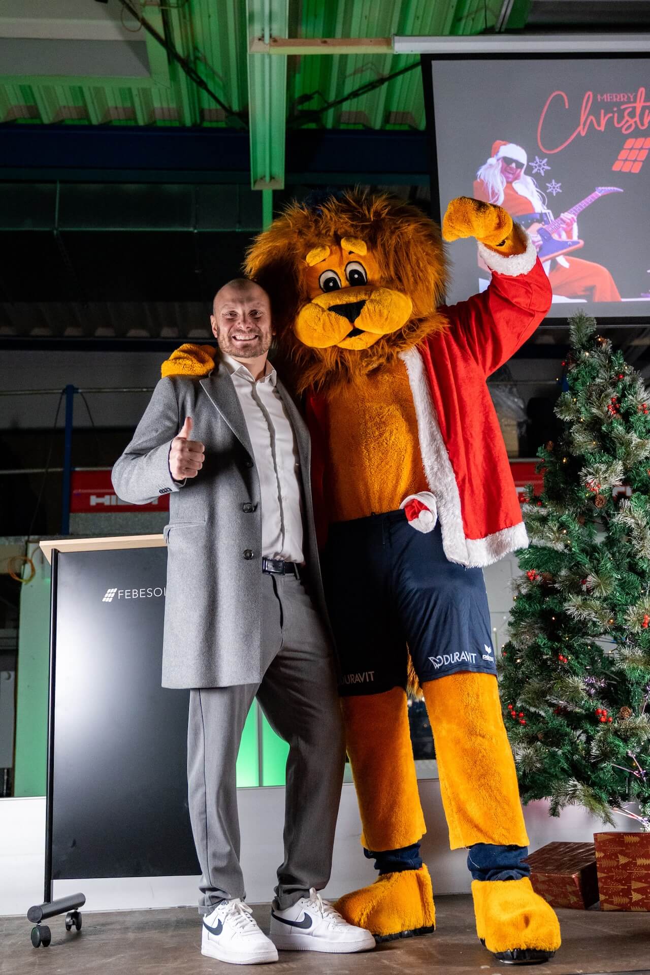 Mann posiert mit Person im Löwenkostüm vor einem Weihnachtsbaum, beide lächeln und strecken den Daumen nach oben.