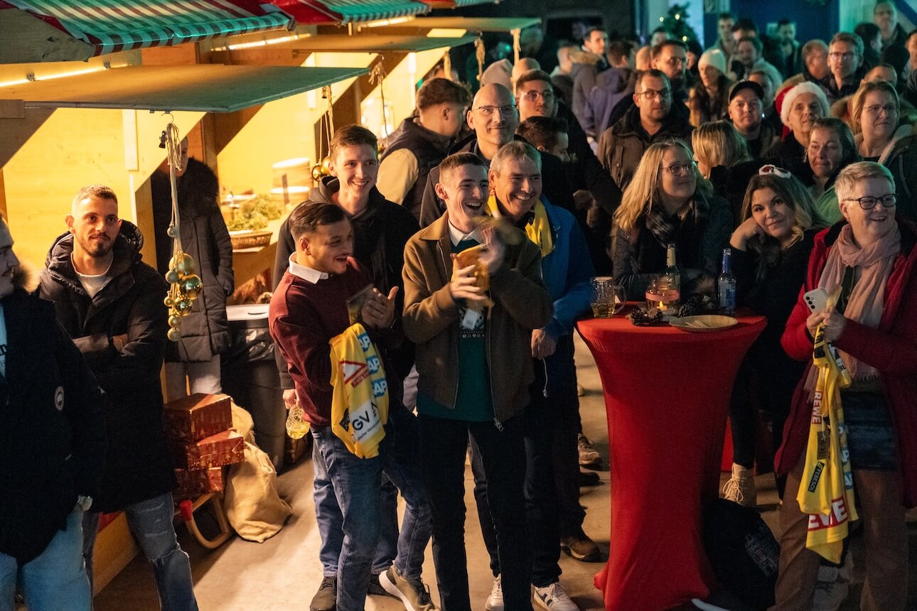 Eine Menschenmenge hatte sich im Innenbereich in der Nähe eines festlich dekorierten Kiosks versammelt, manche mit Getränken in der Hand, andere lächelnd.