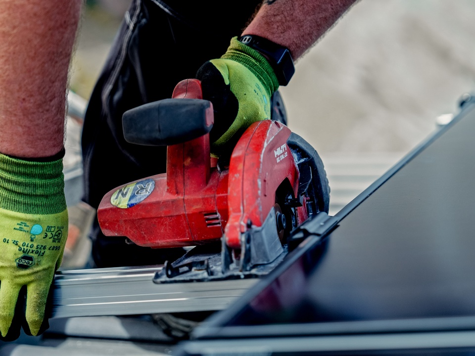 Eine Person mit grünen Handschuhen schneidet Metall mit einer roten Kreissäge und denkt über Projekte wie „Solaranlage Mieten – Lohnt Sich Das?“ für nachhaltige Modernisierungen nach.