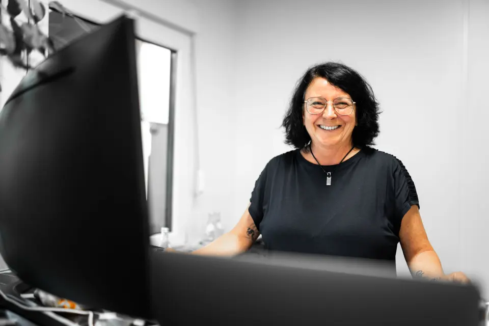 Eine lächelnde Frau mit Brille und dunklem Haar steht in einem Büro hinter einem großen Computermonitor.