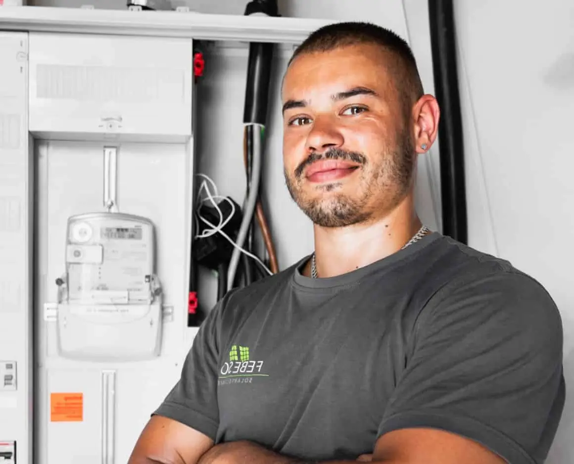 Lächelnder Elektriker mit verschränkten Armen steht neben einem Schaltschrank und trägt ein graues T-Shirt.
