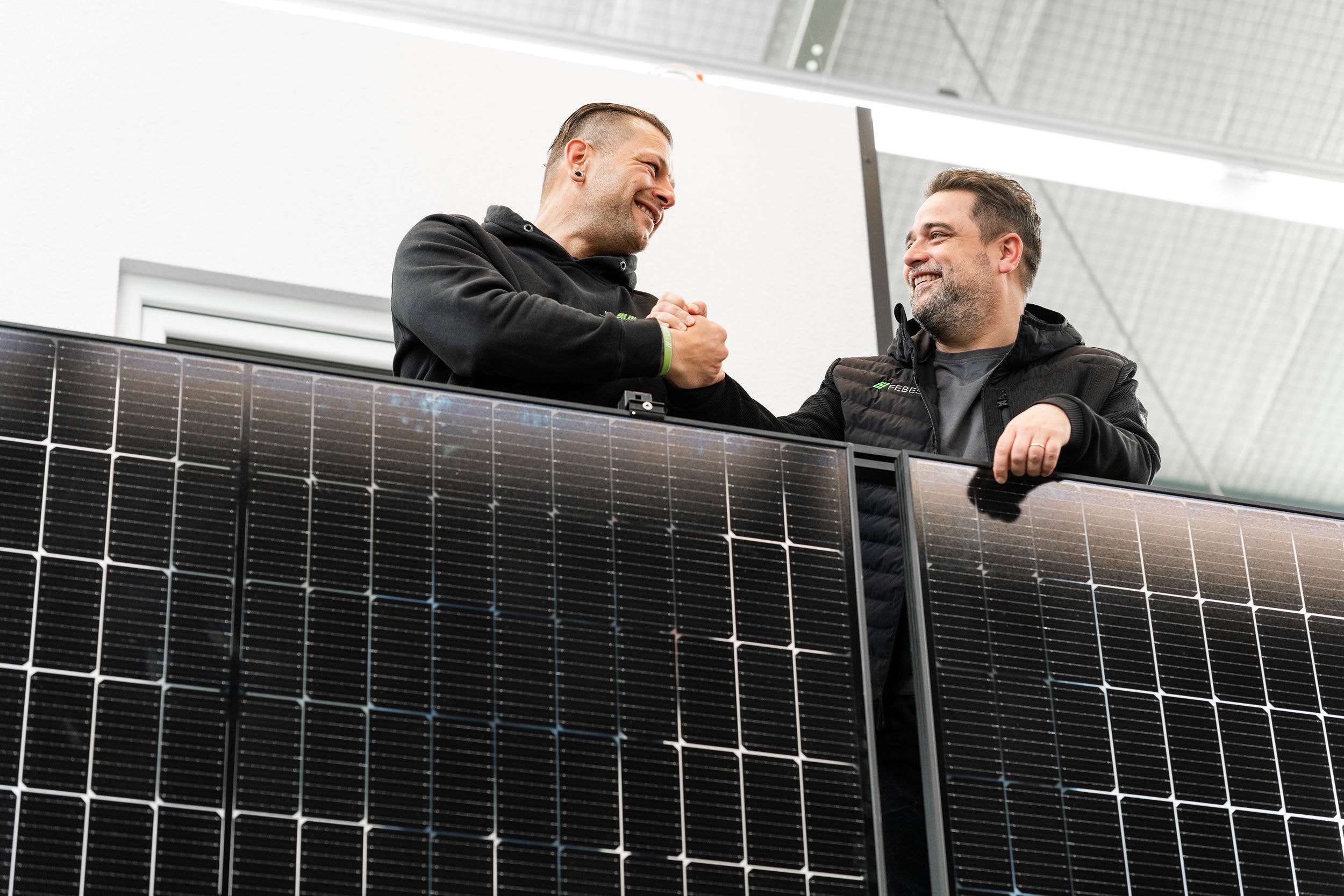 Zwei Männer lächeln und schütteln sich die Hände hinter großen Solarmodulen im Innenbereich und diskutieren über den Abstand zum Nachbarn für eine optimale Installation.