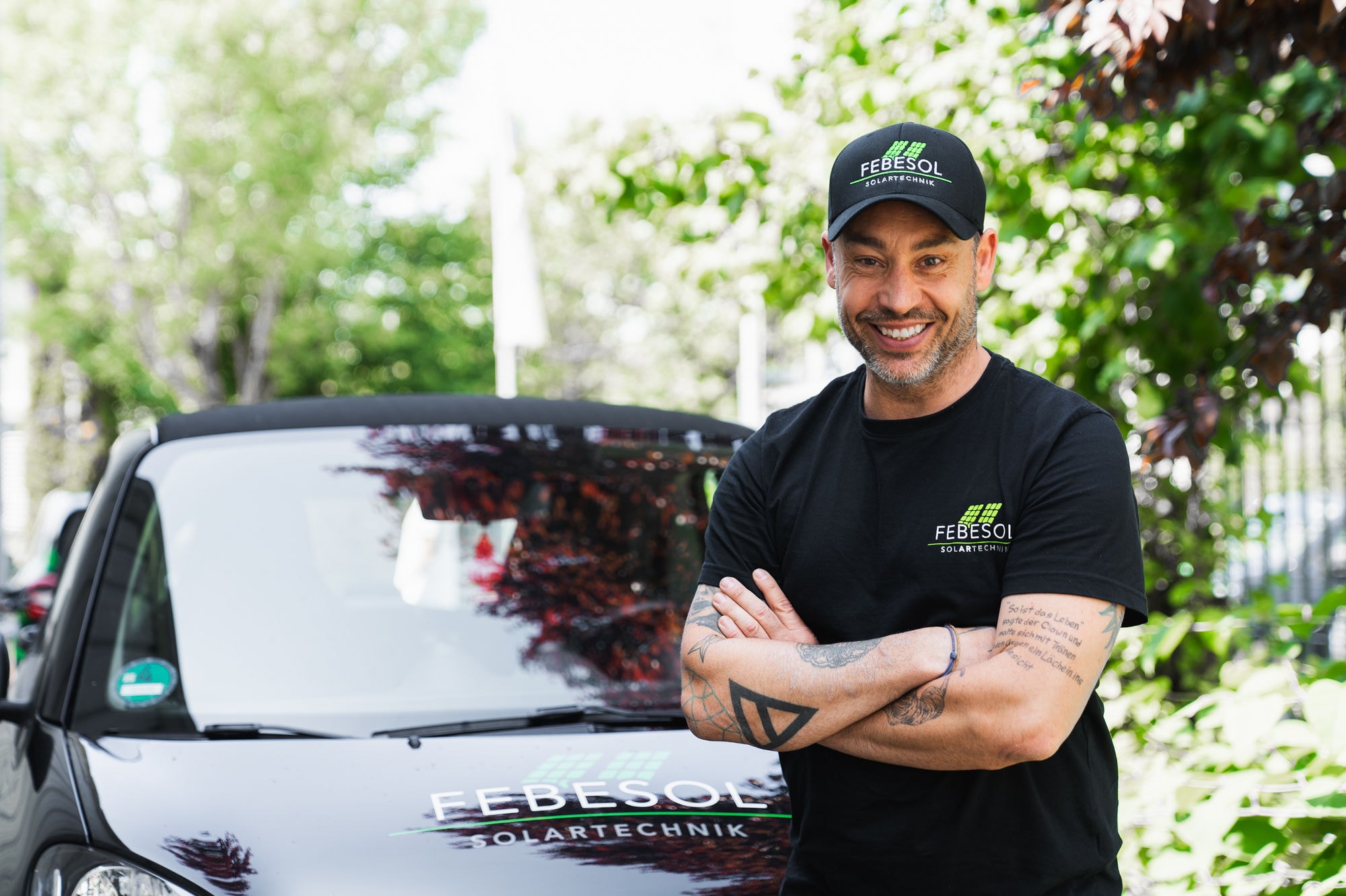 Lächelnder Mann im FEBESOL-Shirt steht mit verschränkten Armen neben einem Auto mit FEBESOL SOLARTECHNIK-Logo und ist bereit zu antworten: „Wieviel Photovoltaik brauche ich für ein Elektroauto?“.