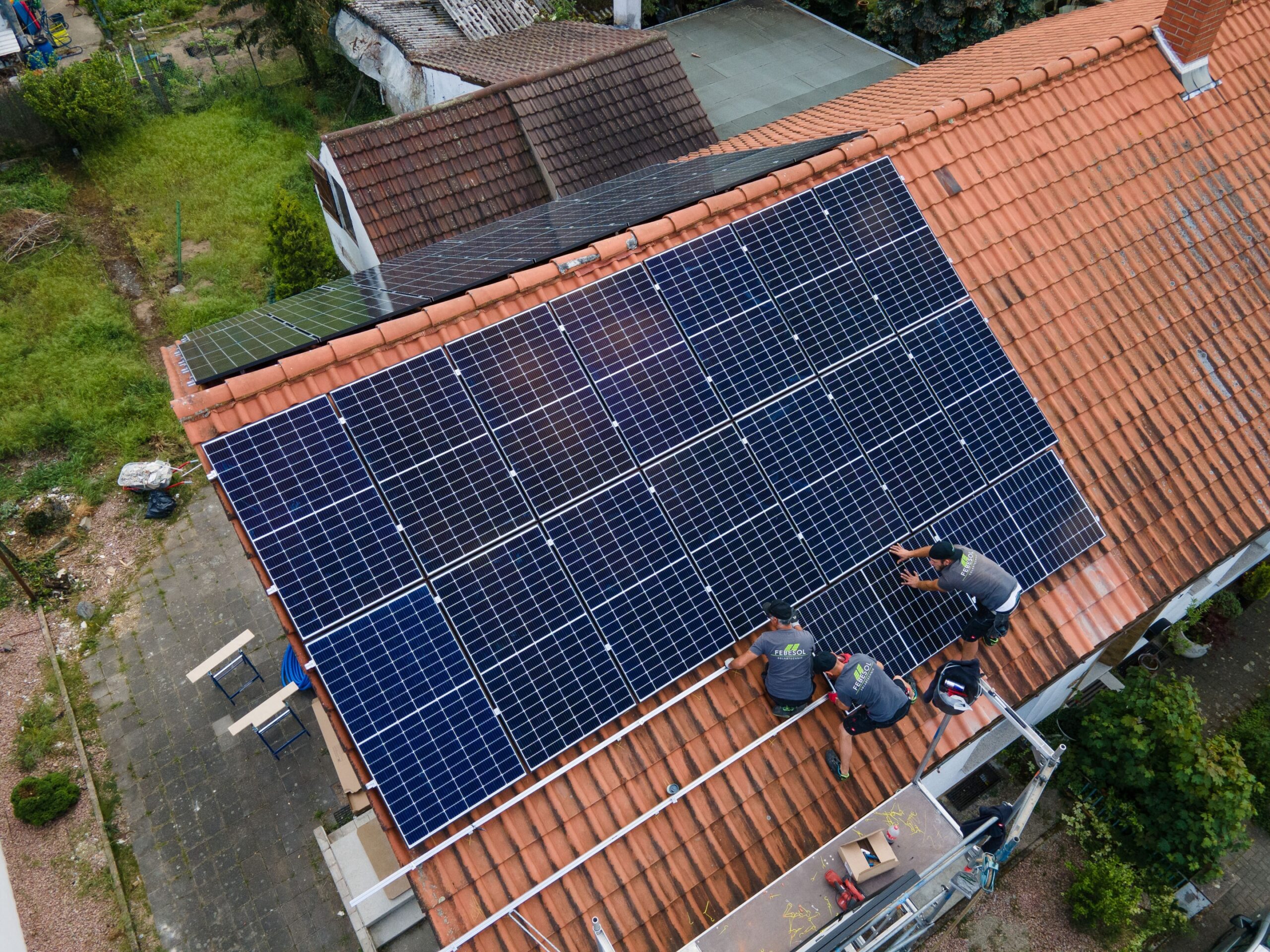 Drei Arbeiter installieren eine Solaranlage vom Typ „Solaranlage kaufen“ auf dem schrägen, rot gedeckten Dach eines Hauses in einem Wohngebiet.