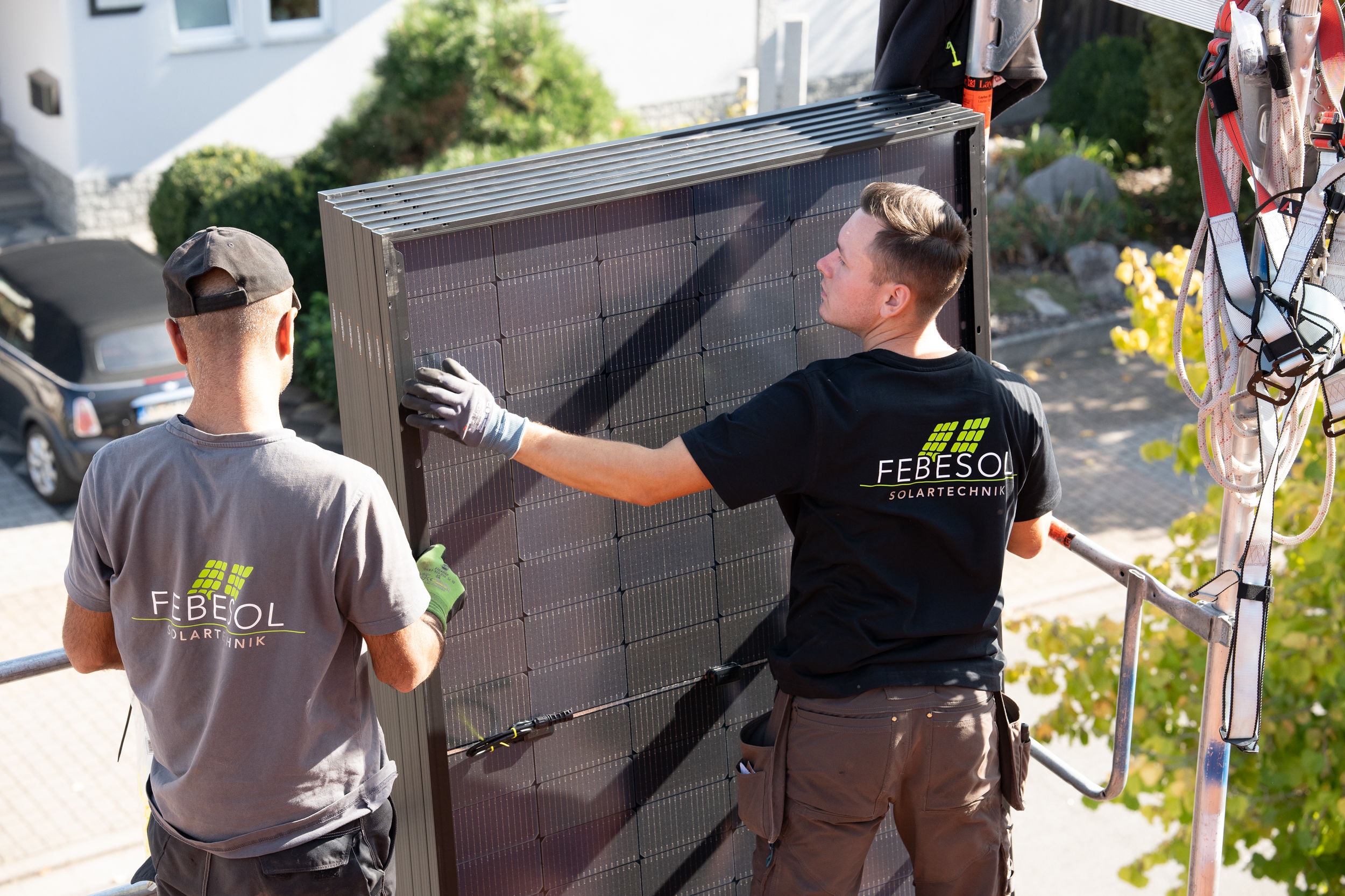 Zwei Arbeiter installieren im Freien ein Solarpanel. Beide tragen Hemden und Handschuhe von „FEBESOL SOLARTECHNIK“ und demonstrieren damit ihr Engagement für Lösungen im Bereich erneuerbare Energien im Rahmen des Solarspitzengesetzes.