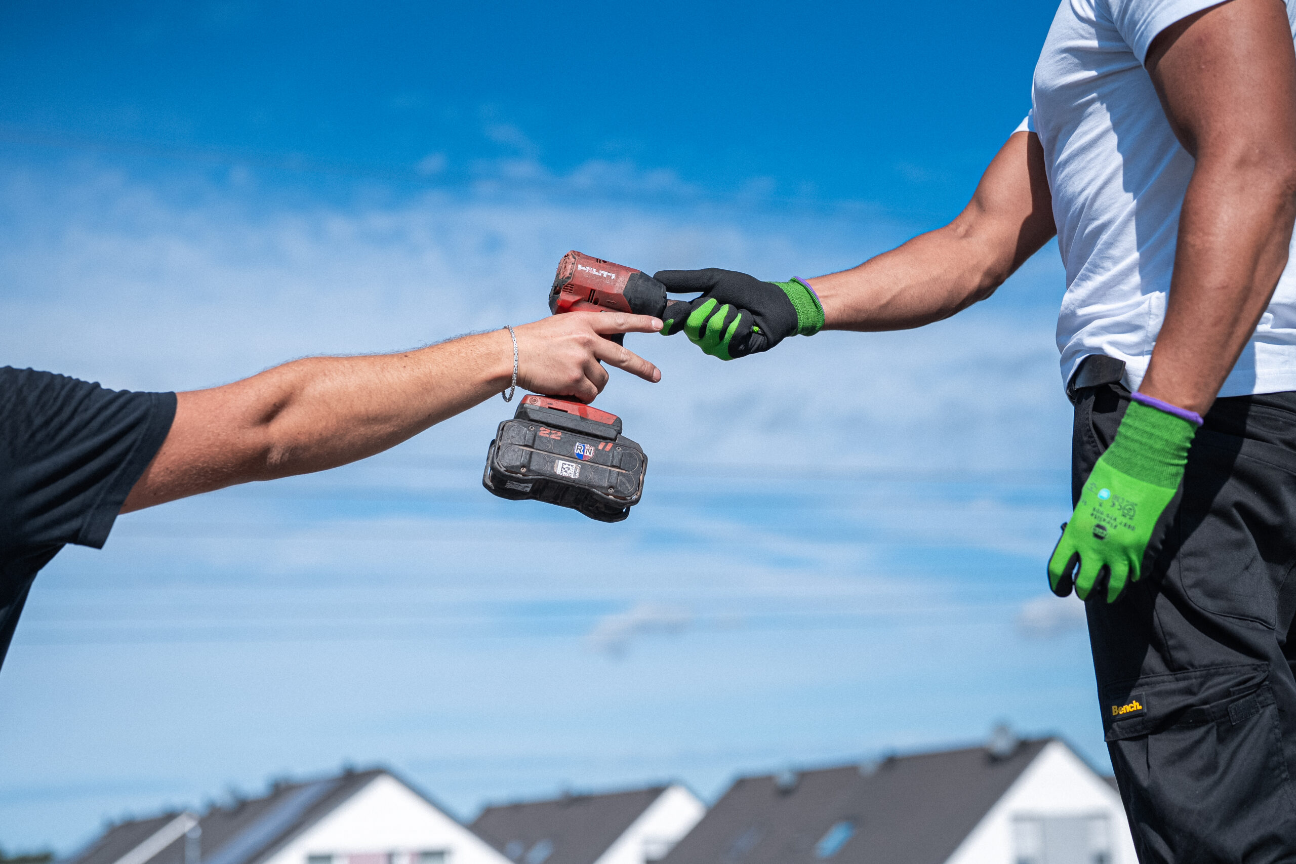 Zwei Arbeiter mit Handschuhen tauschen eine Bohrmaschine im Freien aus, mit Häusern und blauem Himmel im Hintergrund.