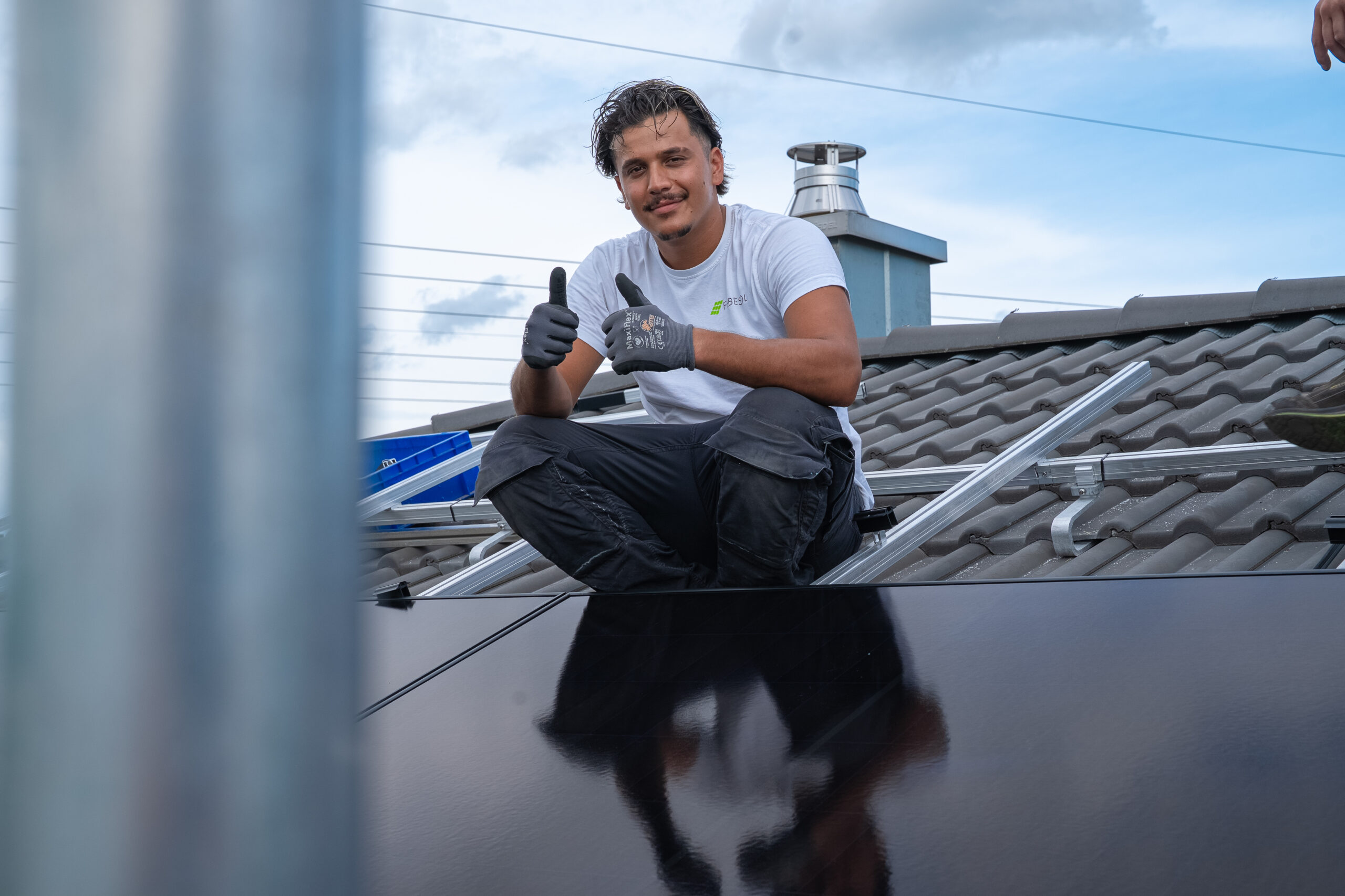 Mann sitzt auf einem Dach neben Solarzellen, lächelt und zeigt zwei Daumen nach oben, mit bewölktem Himmel im Hintergrund.