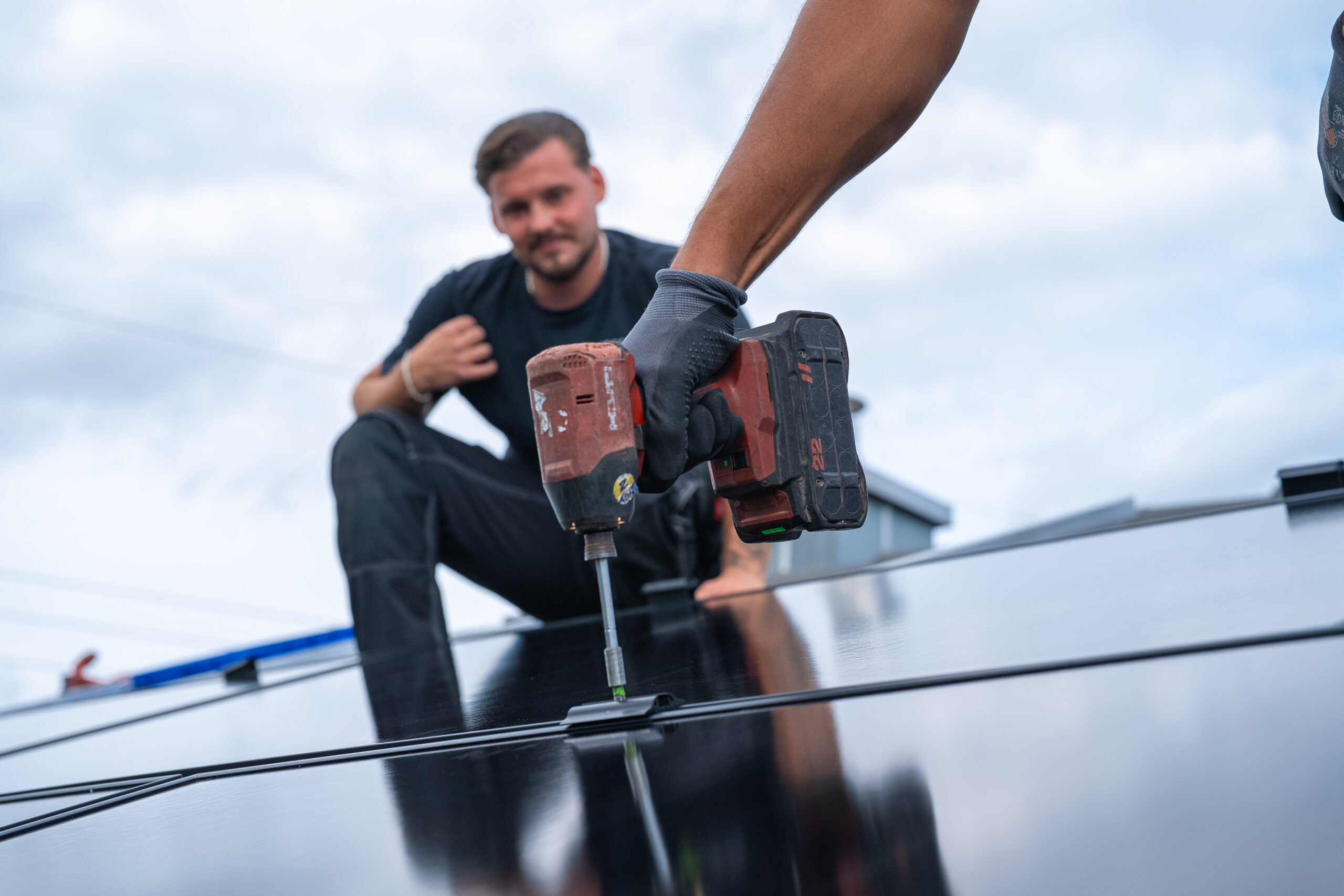 Ein Arbeiter installiert ein Solarmodul mit einer Bohrmaschine, während eine andere Person im Hintergrund auf einem Dach kniet.