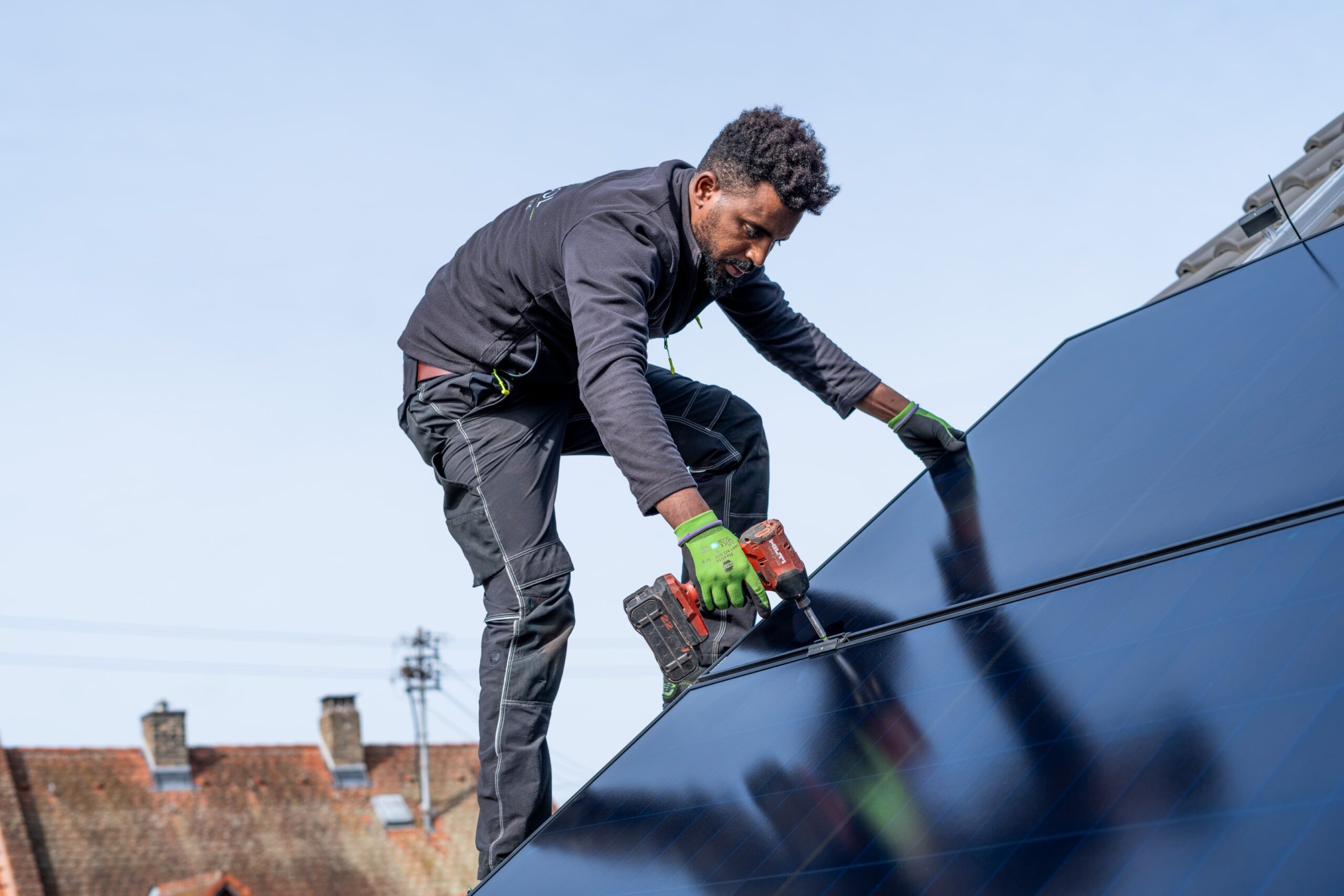 Ein Arbeiter installiert mit Handschuhen und Sicherheitsausrüstung mit einer Bohrmaschine Solarmodule auf einem Dach.