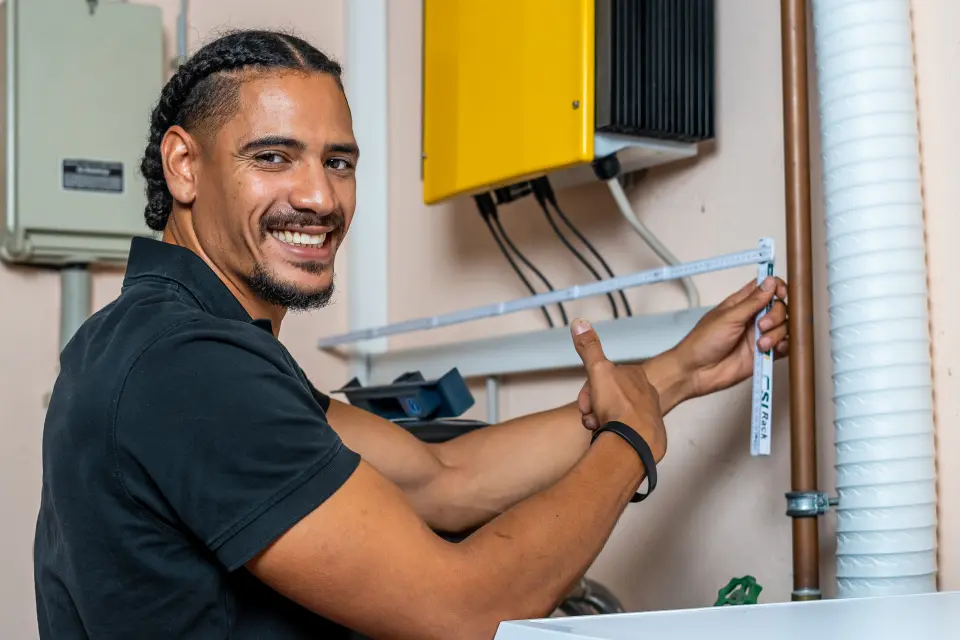 Lächelnder Mann, der die Wandhöhe mit einem Lineal misst und in der Nähe von elektrischen Geräten und Rohren steht.