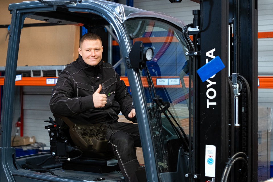 Ein Mann in Arbeitskleidung sitzt auf einem Gabelstapler, lächelt und zeigt den Daumen nach oben in einer Lagerhalle.