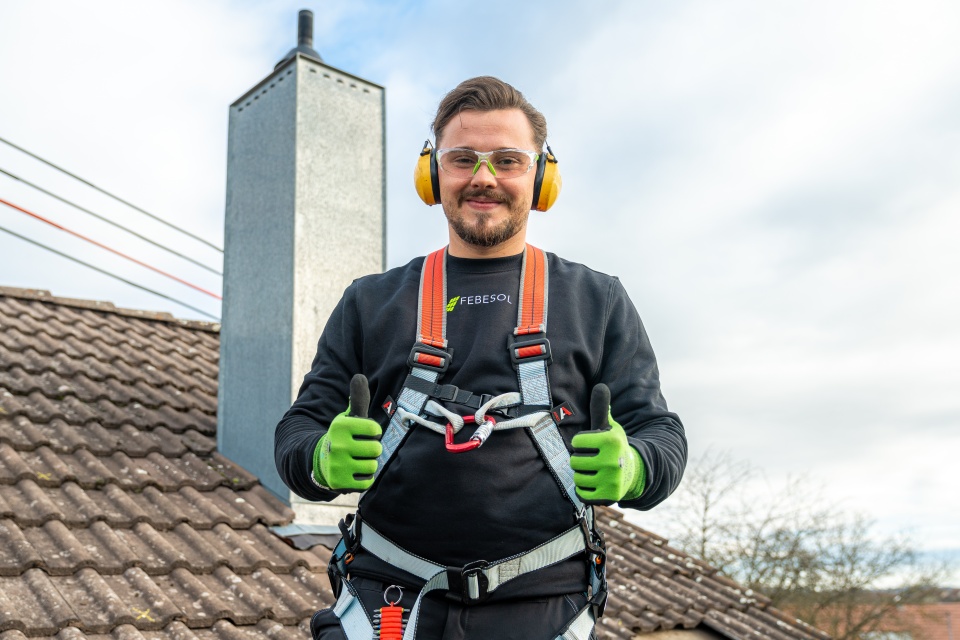 Ein lächelnder Arbeiter in Schutzkleidung steht auf einem Ziegeldach und zeigt zwei Daumen nach oben.