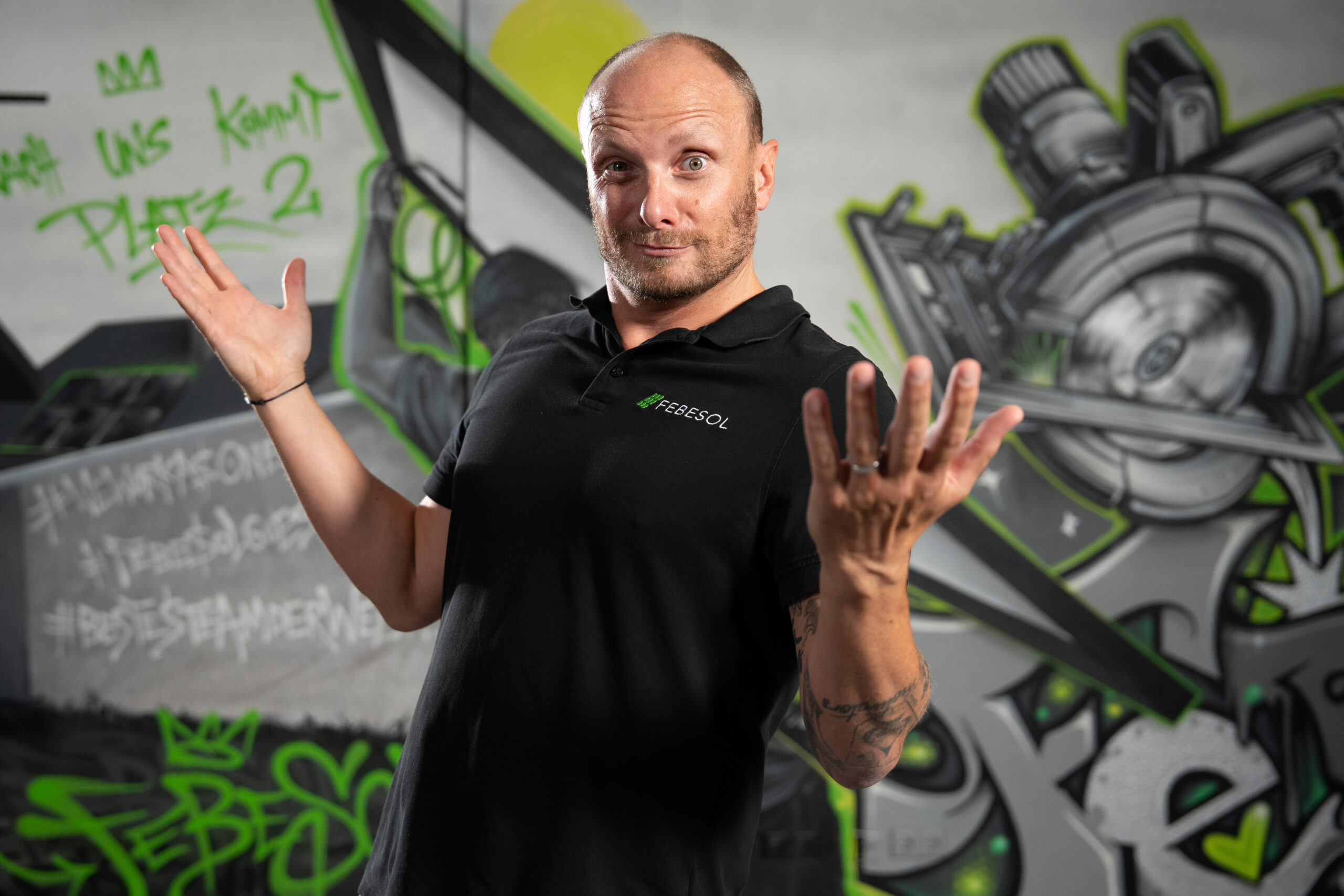 Ein glatzk&ouml;pfiger Mann in einem schwarzen Poloshirt zuckt vor einer bunten Graffiti-Wand.