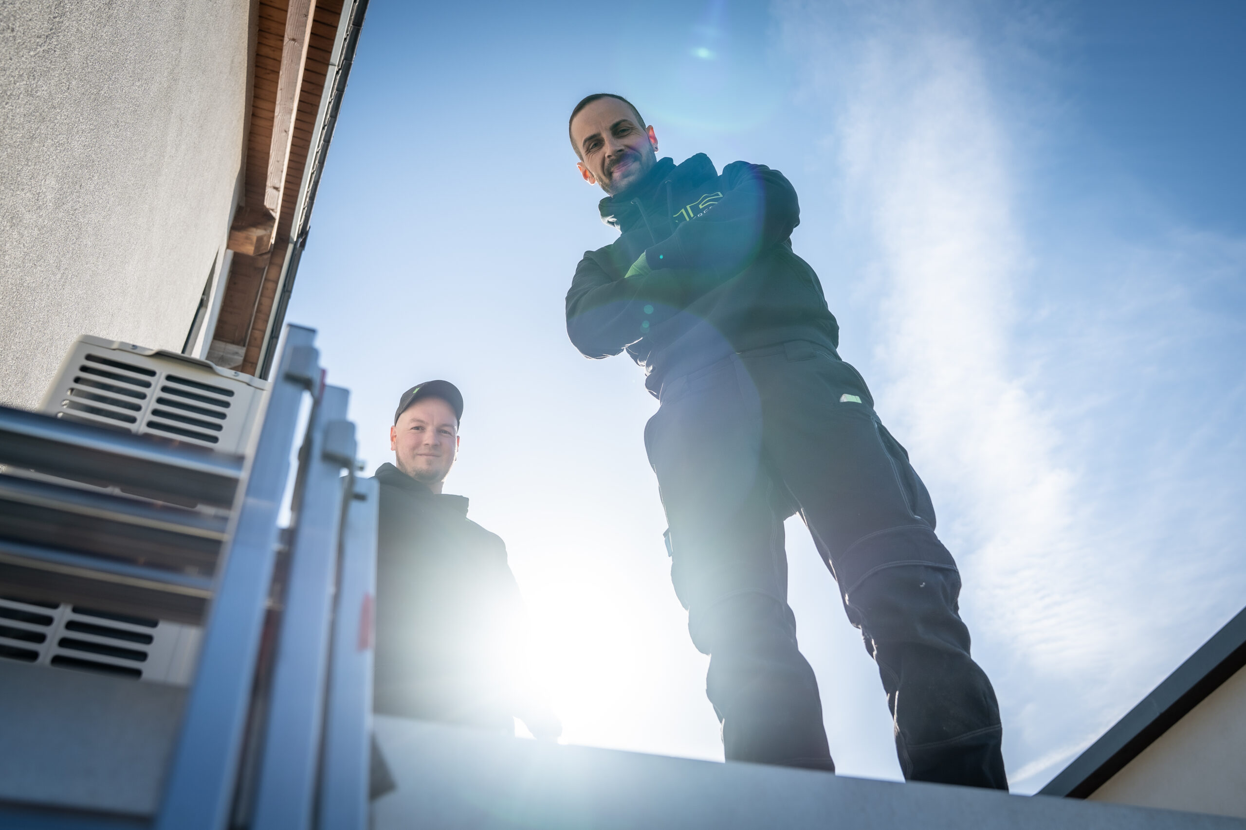 Zwei M&auml;nner in Arbeitskleidung stehen an der Spitze einer Leiter und schauen nach unten, w&auml;hrend die Sonne hinter ihnen scheint.