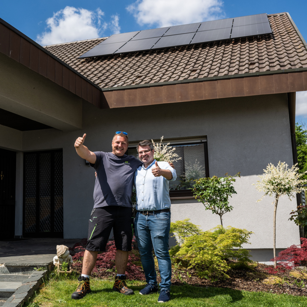 Zwei Männer stehen lächelnd vor einem Haus mit Sonnenkollektoren und zeigen Daumen nach oben in den Garten.